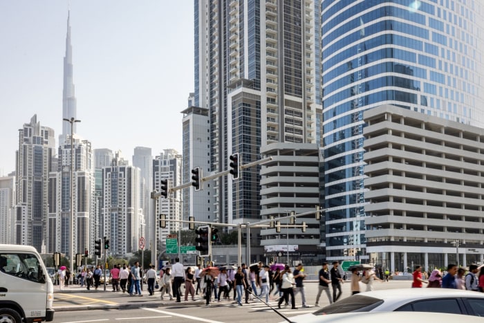 Pedestrians crossing a street in Dubai's Business Bay financial district, with the Burj Khalifa visible in the background.